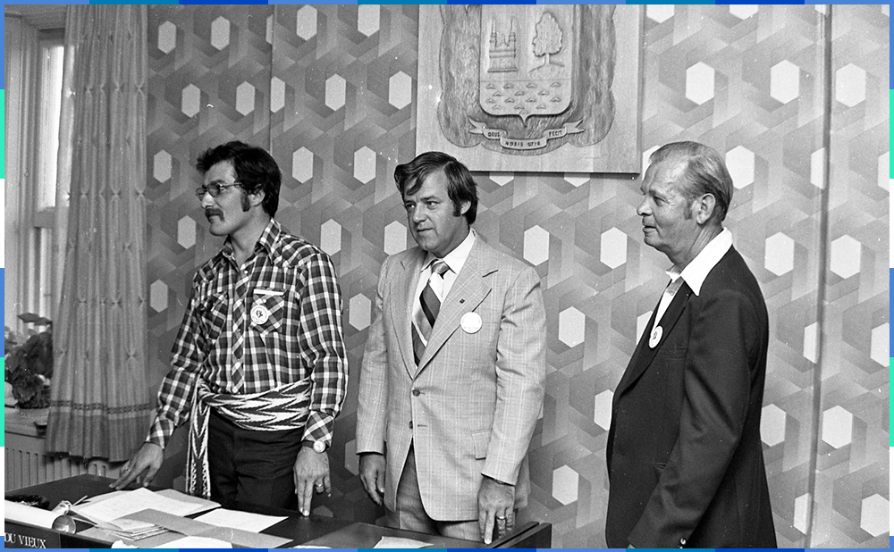 A black and white photograph of three men standing behind a desk. Guy Bélisle and Paul Tardif are wearing suits, while Gilbert Gardner sports an arrow sash. Behind them we can see the coat of arms of the City of Saint-Eustache.
