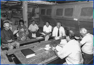 The eight board members are sitting around a wooden table inside the Légaré mill. On the right side of the photo, we can see the bolting cloth used to separate the flour from the bran.