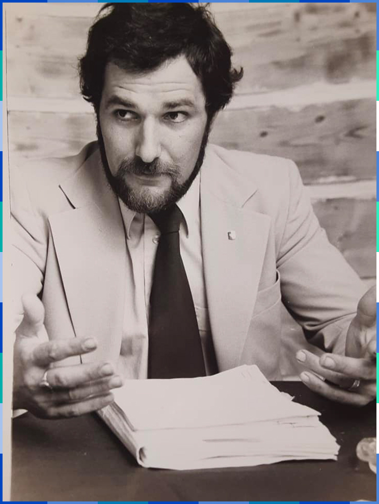 A black and white photograph of a man with a beard wearing a suit and tie. He is sitting at a desk with a pile of documents in front of him.