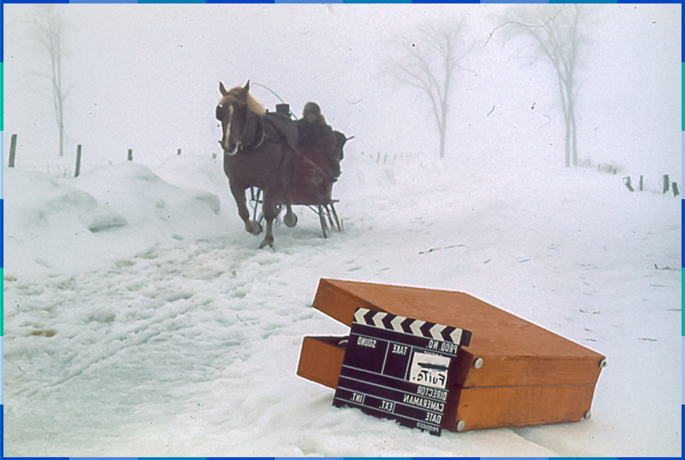 A colour photograph of a scene from a film that takes place outdoors in winter. In the foreground there is a suitcase on the snow, which is being used to hold a clapperboard. The scene features two men sitting on a red sleigh pulled by a brown horse.