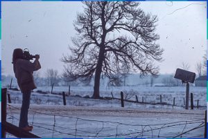 The photo was taken outdoors in winter. On the left, we see a camera operator standing. In the middle of the photo is a tall tree in the middle of a field. On the right is a letterbox, which appears to be damaged.