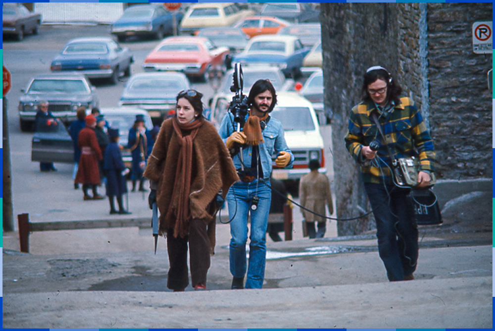 In the middle of the image, a woman and two men are walking up a sloping paved street. The men are moving a movie camera and audio equipment. Behind them are around twenty parked cars, and actors in costumes.