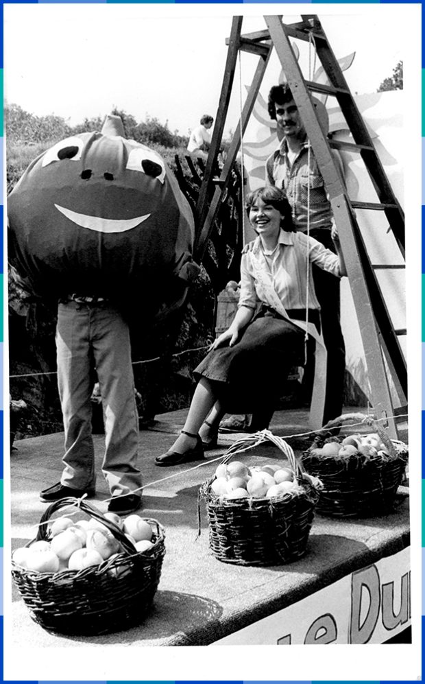 A black and white photograph of a man standing and a woman sitting under a stepladder having their photo taken with a person dressed up as a smiling apple. In front of them are three baskets full of apples.