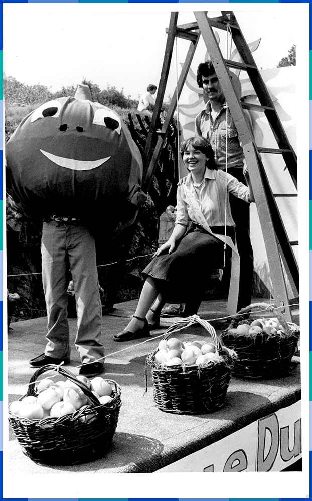 A black and white photograph of a man standing and a woman sitting under a stepladder having their photo taken with a person dressed up as a smiling apple. In front of them are three baskets full of apples.