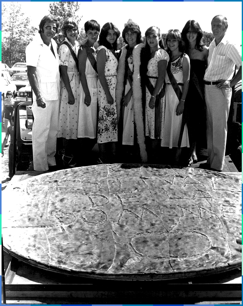 A black and white photograph of seven young women and two men posing behind a huge apple pie on which the words “Festival de la pomme” (“Apple Festival”) are written.