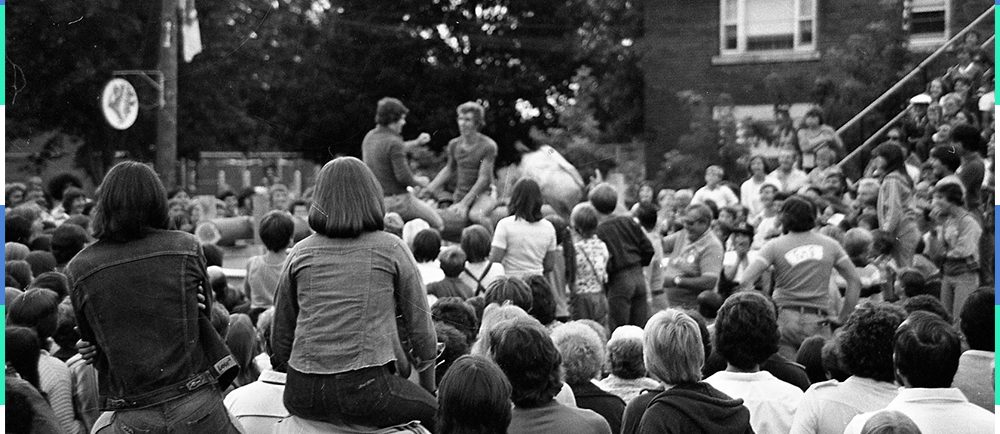 A black and white photograph of a crowd gathered in the street around two men who are sitting on a raised platform. Two girls are sitting on another person’s shoulders.