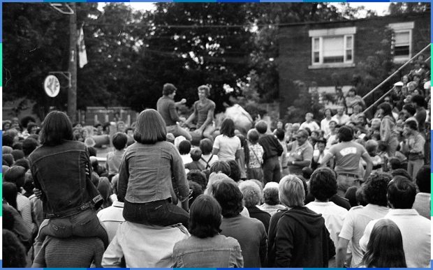 A black and white photograph of a crowd gathered in the street around two men who are sitting on a raised platform. Two girls are sitting on another person’s shoulders.
