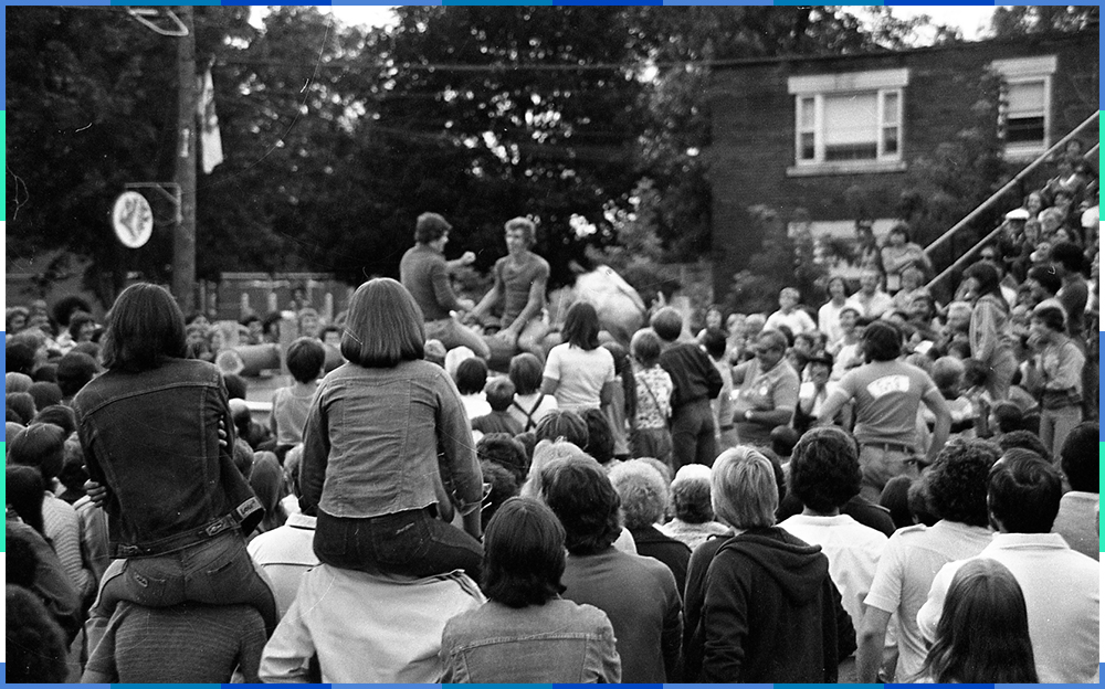 A black and white photograph of a crowd gathered in the street around two men who are sitting on a raised platform. Two girls are sitting on another person’s shoulders.