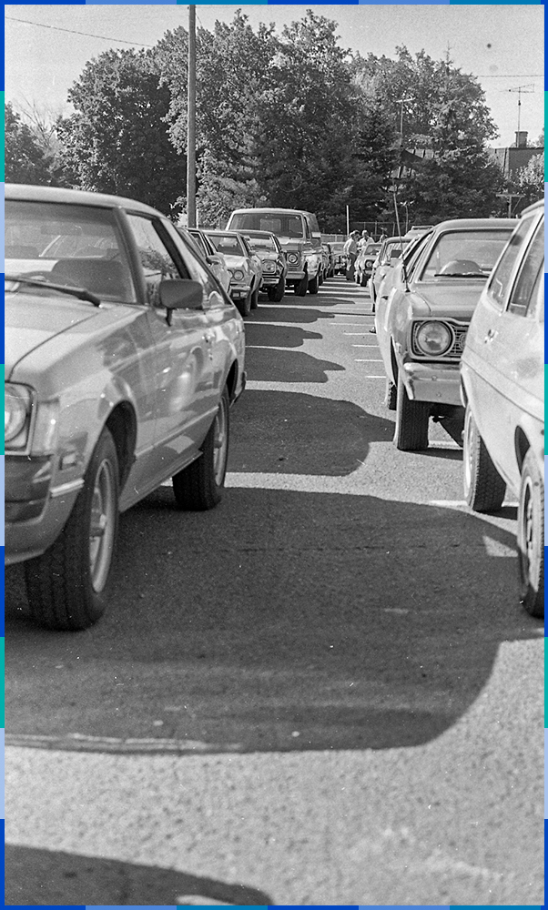 Two rows of cars, parked side by side, stretch as far as the eyes can see. In the distance, we can see a group of men talking.