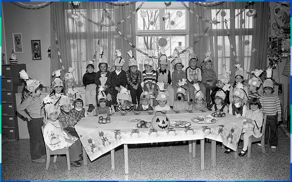 Around thirty children who appear to be about ten years old are gathered around a table in a room decorated for Halloween. They are all wearing festive hats.