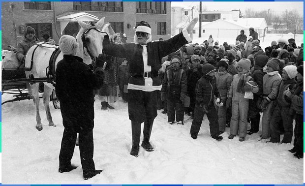 A black and white photograph of a man dressed up as Santa Claus visiting children on the grounds of an elementary school. His presents are in a carriage pulled by a white horse.