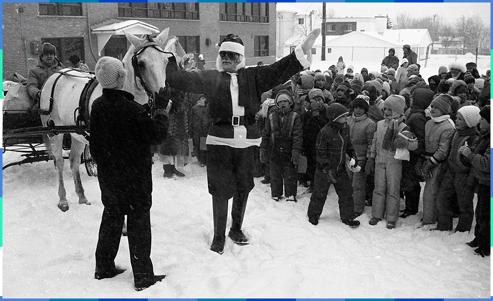 A black and white photograph of a man dressed up as Santa Claus visiting children on the grounds of an elementary school. His presents are in a carriage pulled by a white horse.