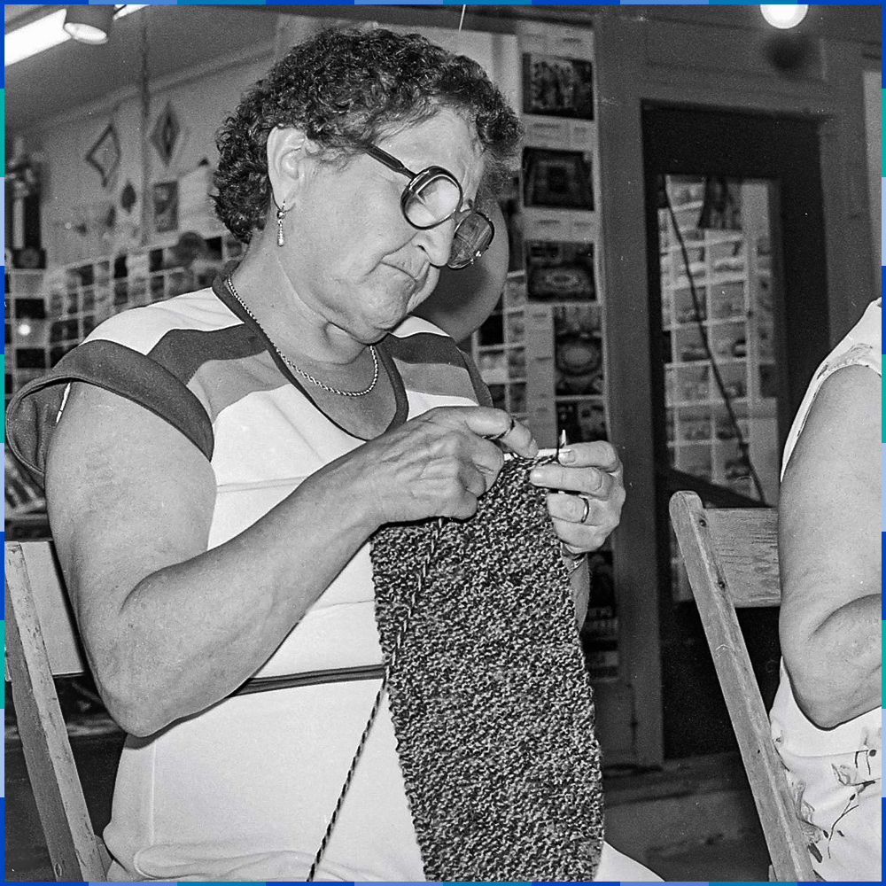 A waist-up portrait of a woman with short, curly hair, wearing glasses. She is seated and knitting, her eyes focused on the needles.