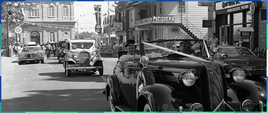 A black and white photograph of cars driving down Rue Saint-Eustache. Shops can be seen on the right side of the street, and the Saint-Eustache church is in the background.