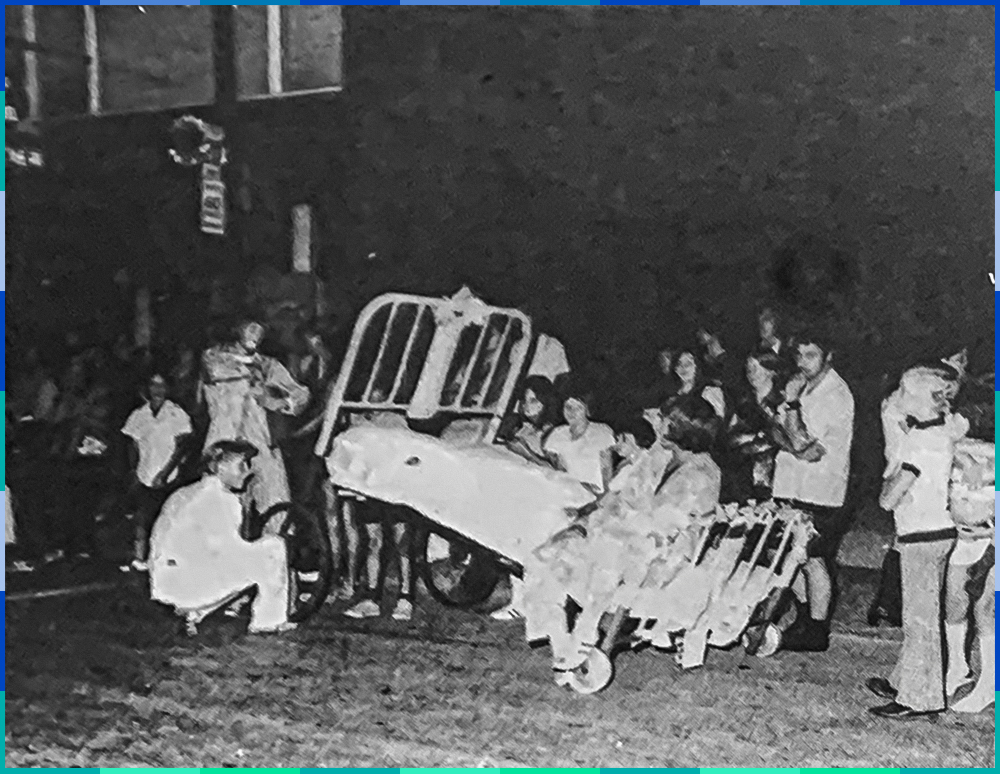 In the middle of the black and white photograph is a hospital bed, with metal bars, on wheels. One person is sitting while another appears to be adjusting a wheel. A small crowd is watching them.