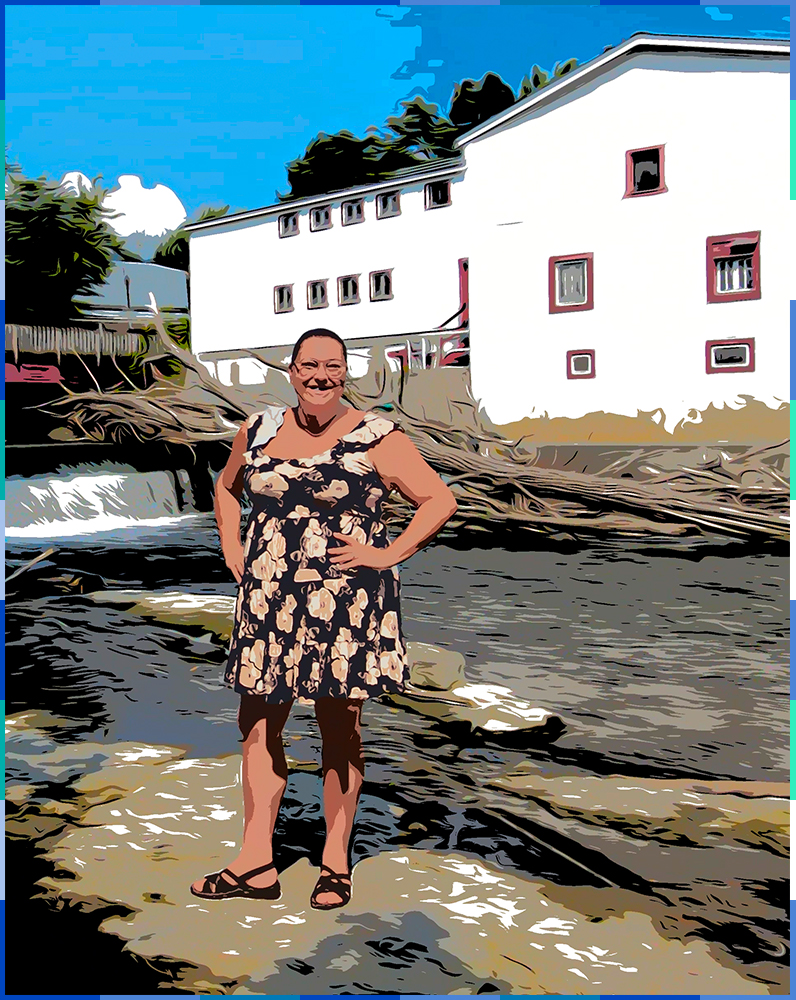 A photograph of a smiling, brown-haired woman wearing glasses and a floral print dress, digitally edited to look like a comic strip. She is standing on a large, flat rock, and has her hands on her hips. Behind her are the Légaré mill, the dike, and the Rivière du Chêne.