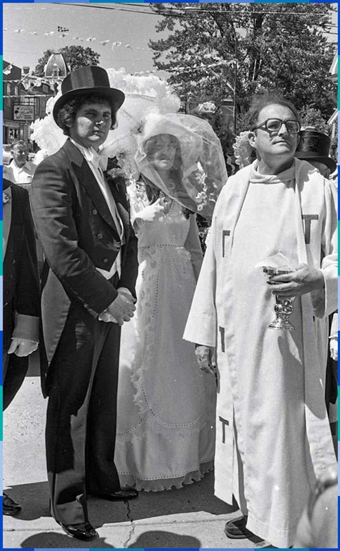 Black and white photograph taken outdoors of a couple dressed in wedding attire. The man wears a tailcoat and a top hat while the woman wears a dress and a hat decorated with a veil. A priest in a cassock is at their side.