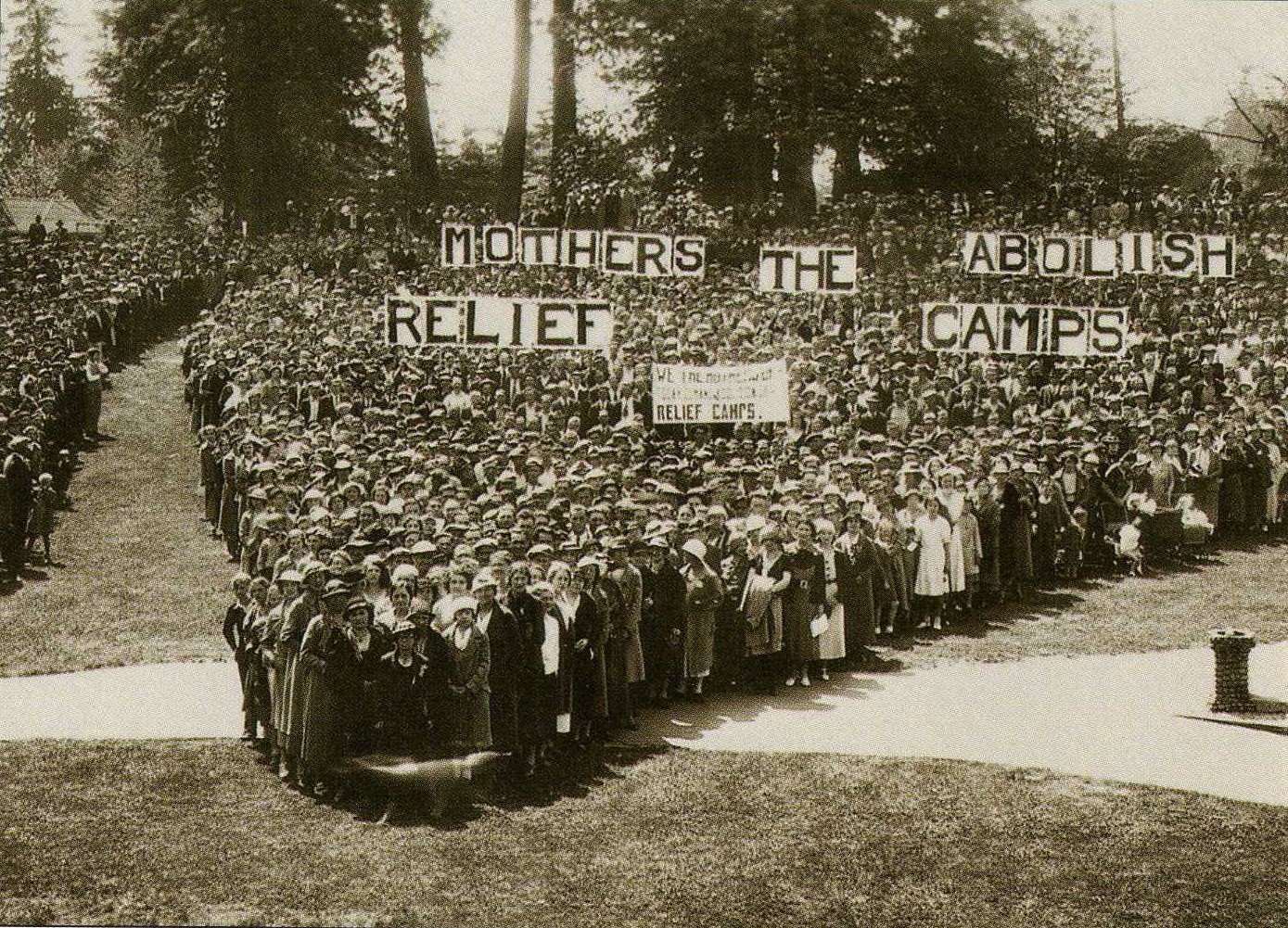A black and white photograph, from a bird’s eye view of a large group of female protesters gathered together, carrying letter banners that spell out, Mothers Abolish The Relief Camps.