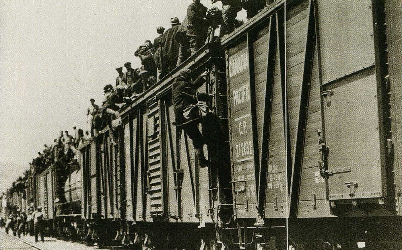 A black and white photograph of workers standing on top of a Canadian Pacific train with others standing by the tracks attempting to climb on the train. The focal point of the photo is of a man climbing up the train to join the group standing on top.