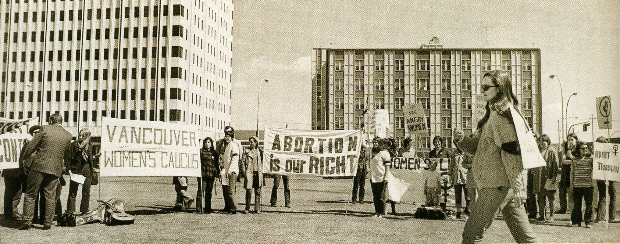 A black and white photograph of a group of protesting women standing in front of two buildings, carrying signs and banners with one stating Abortion Is Our Right and another one that states Vancouver Women’s Caucus. One woman walks across the space in front of the group and on the left, a man in a suit speaks to a person in the crowd.