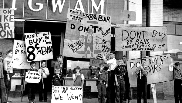 Black and white photograph of a group of children standing outside a store called WigWam, some licking ice cream cones, holding placards saying variations on the wording ‘Don’t be a Sucker, Don’t buy 8c bars’.