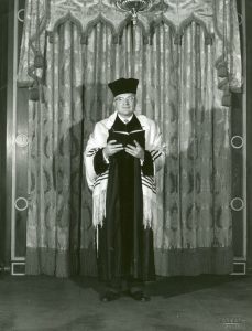 A black-and-white photograph of Cantor Nathan Mendelson in front of a synagogue ark. He is wearing a black cantor's hat, black robes, and a tallit, and is holding a prayer book.