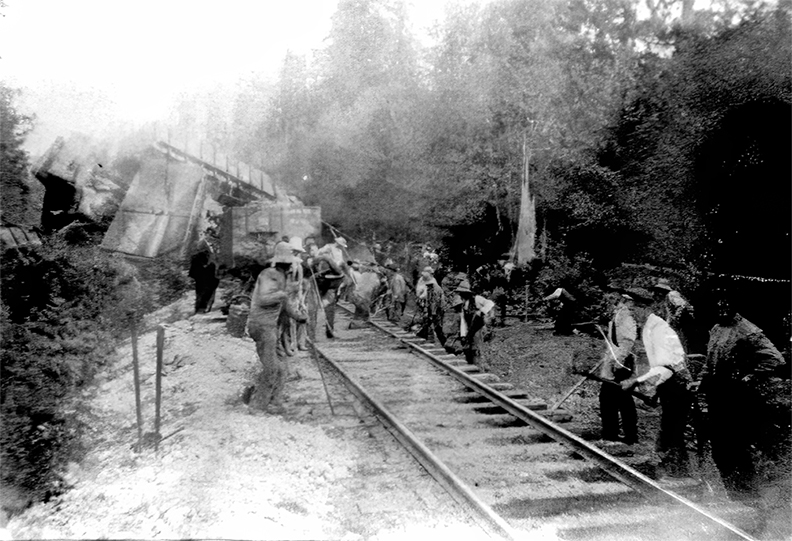 Black and white photograph of a railway track in the foreground along which groups of men, many of which are Chinese, are working. In the background is the wreckage of numerous wooden railway coal cars ‘concertina’d’ together in a jumbled pile.