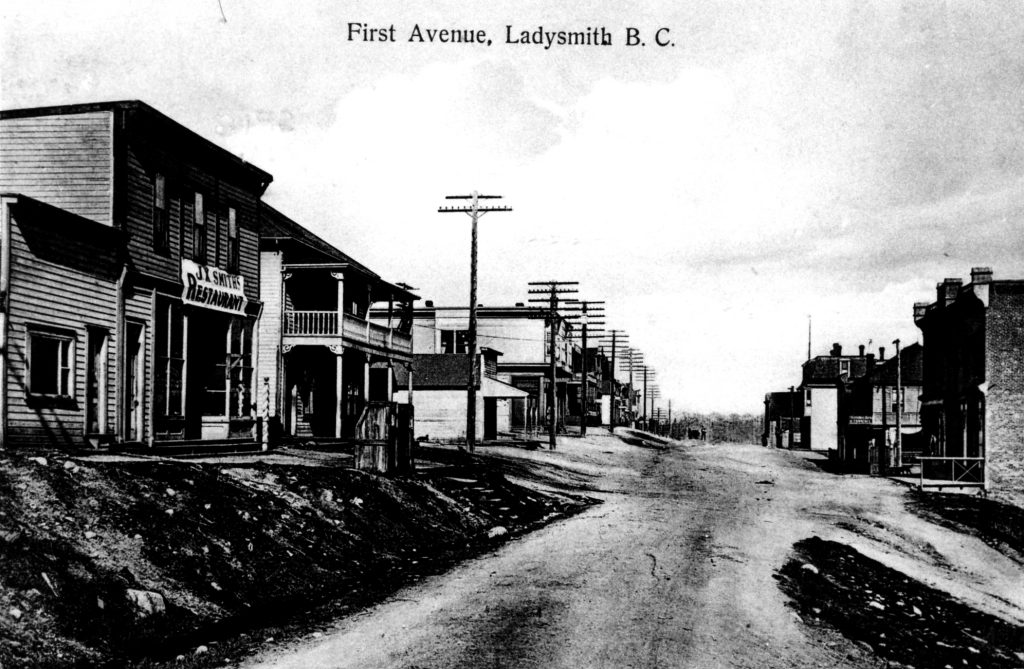 Black and white photograph of an unpaved town street flanked by a variety of wooden and brick buildings. At at centre-left is a two story clapboard sided flat roofed building with a large windowed shop front over which is a canvas sign with the lettering ‘J.X. Smith’s Restaurant’.