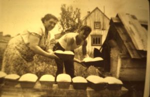 Sepia photograph: a woman and a man remove bread molds from an outdoor oven. In front of the oven: other bread moulds. Background: wooden house and vegetation.