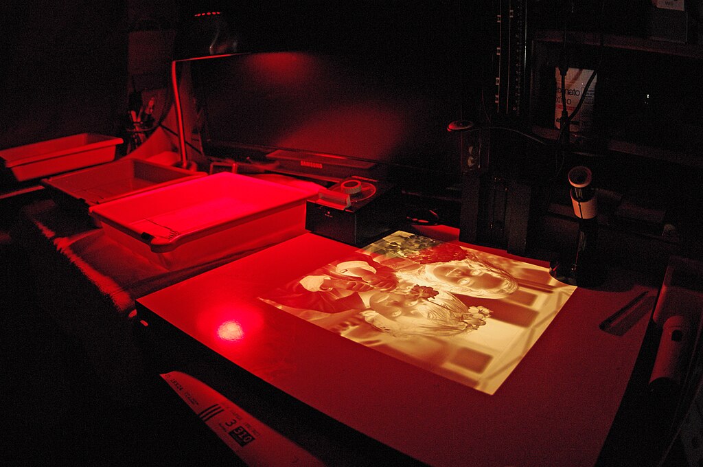 A photographic darkroom illuminated by a pale red light. Visible in the photo are several trays holding liquids. Exposed and drying photographs are on the table beside them.