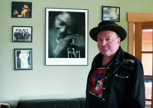Colour photograph of man dressed in black shirt jacket over a colourful T shirt with black brimmed hat standing in front of a light wall adorned with framed photographs of people, some with musical instruments.