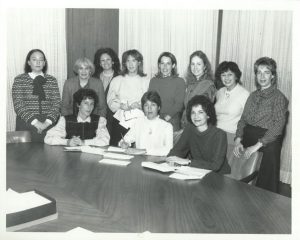 A black-and-white photograph of a group of women standing around a table.
