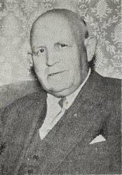 Black-and-white portrait of a man dressed in a dark three-piece suit with a light-colored shirt and tie, seated in front of a patterned wallpaper. He has a calm expression and appears to be in later life.