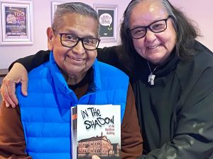 Colour photograph showing a First Nation man holding a book, with a woman holding her arm around his shoulders.