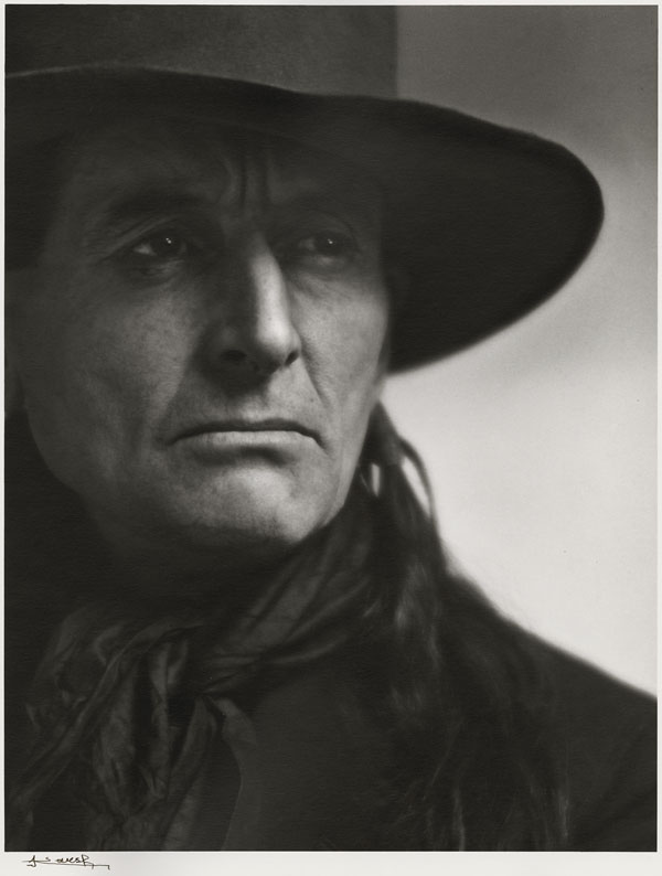 Black and white portrait of Grey Owl. The portrait is a close-up of the man's face. Framed to the right, Grey Owl's face and gaze are turned slightly to the right. Dressed in black, he is wearing a cowboy hat, a scarf and a jacket. A lock of hair falls over his shoulder. The photograph is signed Yousuf Karsh.