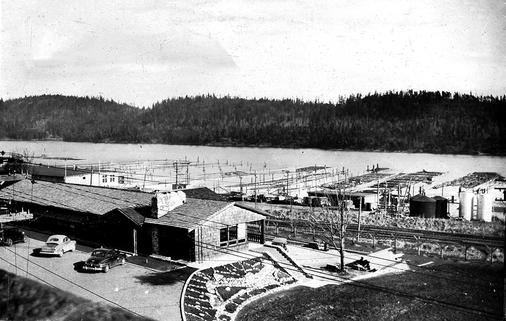 Black and white photograph of a single-story office building with several cars parked out front. Ordered log booms float in the harbour waters in the background.