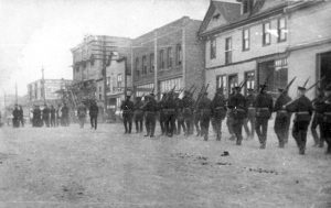 Black and white photograph of soldiers, rifles over their shoulders, marching along an unpaved town street.