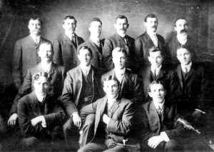 Black and white photograph of a group of men dressed in three piece suits, seated arranged in three rows.