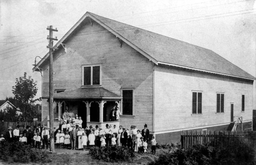 Black and white photograph of a single-storey gable-roofed simple rectangular wooden building with front porch about which a group of well dressed men, women and children are standing facing the photographer.