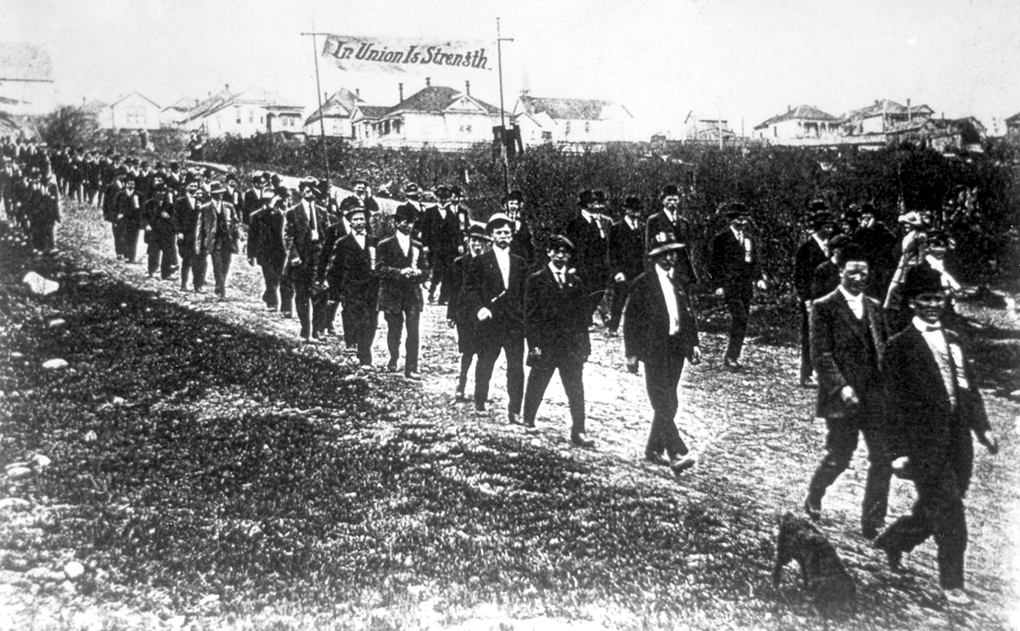 Black and white photograph of men in civilian clothes parading downhill along an unpaved street carrying a banner with the wording ‘In Union Is Strength’. Some men wear prominent lapel pins.