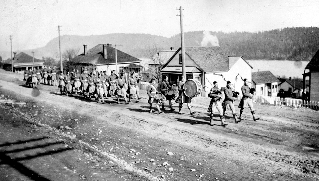 Black and white photograph of a formation of kilted soldiers led by a band consisting of three bagpipers and three drummers marching along an unpaved street lined by detached wooden houses, between which can be glimpsed the waters of a harbour.
