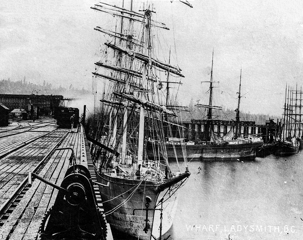 Black and white photograph of four ocean-going sailing vessels, sails furled, tied alongside two large wooden wharves.
