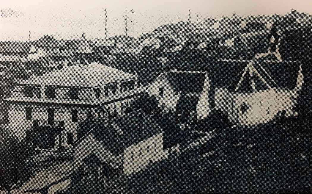 Black and white panoramic view of a town with a cluster of four buildings in the foreground, one of which has characteristic church architecture.
