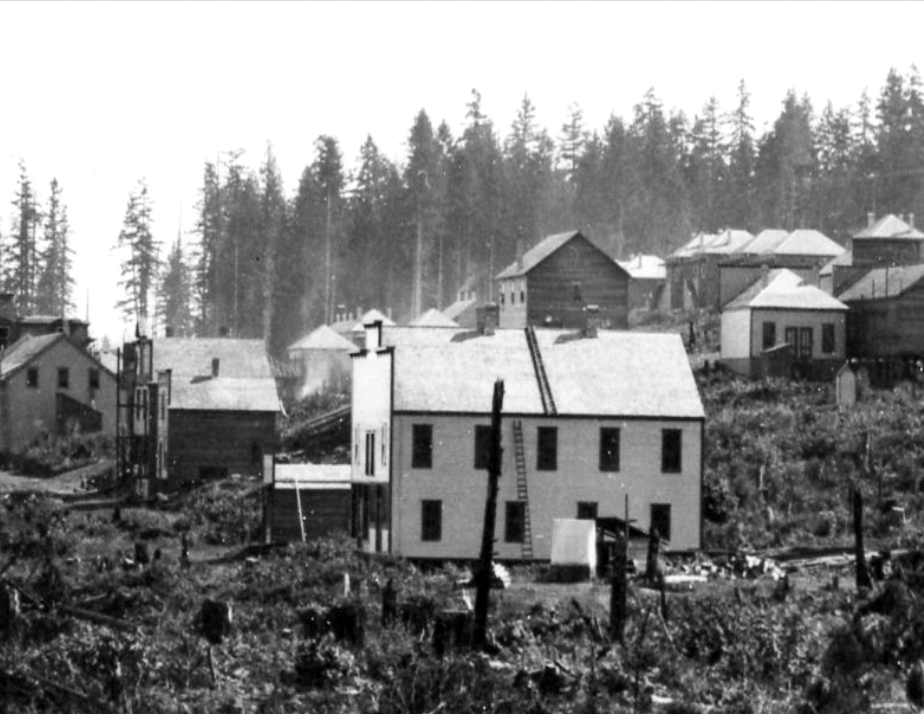 Black and white photograph of a light coloured two-storey wooden rectangular building with pitched roof and large rectangular windows staggered between levels surrounded by open space occupied by scrub bushes and tree stumps. Other wooden buildings of similar construction are in the background.
