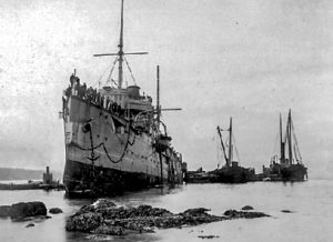 Black and white photograph shows oblique port side waterline bow view of an iron hulled battleship aground on rocks. Crew members line the deck railings. Several smaller vessels are attending.