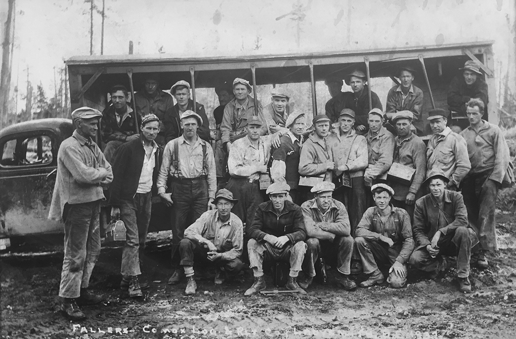 Black and white photograph of a group of 26 men in work clothes, some carrying lunch buckets, posed in front of a truck modified as a crude open-sided bus.