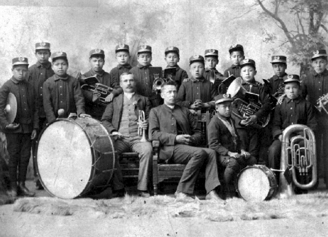 Posed black and white photograph of 15 First Nation boys in capped simple uniform carrying brass band instruments standing behind two Caucasian seated men, one of whom is a priest.