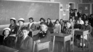 Black and white photo showing neatly dressed students sitting at their desks in a classroom.