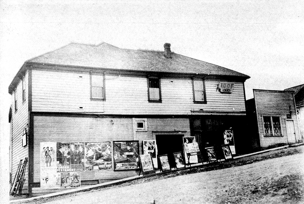 Black and white photograph of a wooden two-storied building viewed across a steeply sloping unpaved street. Numerous billboards with advertisements adorn the walls at street level.