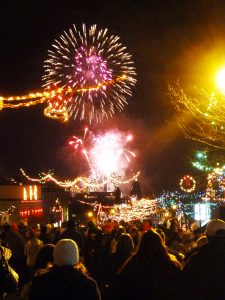 A night time colour photograph showing a town street adorned with colourful Christmas lights packed with people watching an overhead fireworks display.